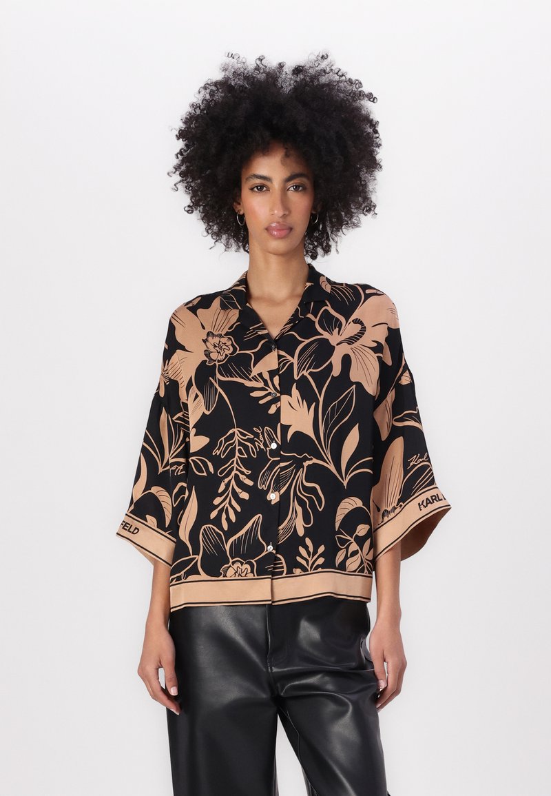 Sort blomstret bluse med beige botanisk print, korte ærmer, løs pasform og knaplukning. Inkluderer et brandlogo langs sømmen.