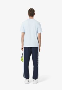 Lichtblauwe t-shirt met korte mouwen, gecombineerd met marineblauwe trainingsbroek met een zijlogo en witte accenten. Iemand houdt een mesh tas met tennisballen vast.