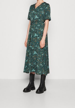 Kvinde iført en grøn midi-kjole med korte ærmer og sorte chunky ankelstøvler, stående mod en ensfarvet lys baggrund.