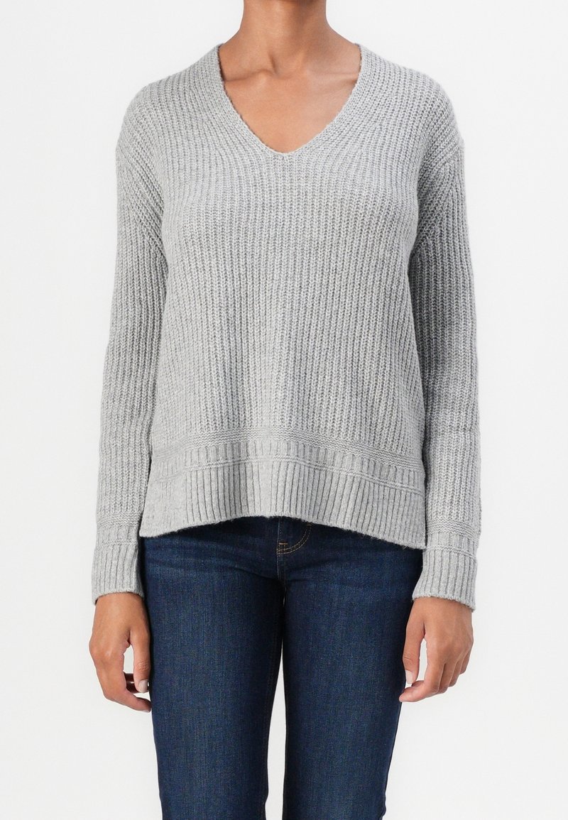 Femme portant un pull en maille côtelée gris clair à col en V avec manches longues et un jean bleu foncé, debout devant un fond blanc uni.