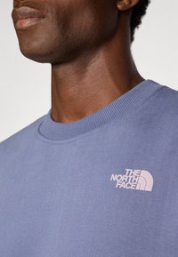 Nærbilde av en person som har på seg en blå genser med rund hals, med den rosa The North Face-logoen på brystet.