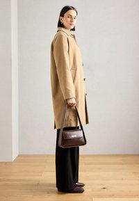 Manteau camel avec un devant boutonné, associé à un pantalon noir évasé et des bottes à talons marron, tenant un sac à main en cuir marron.