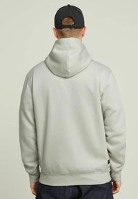 Haut à capuche gris clair avec cordon de serrage, poignets et taille côtelés. Présente une petite étiquette de logo sur le côté inférieur, en tissu de coton doux.