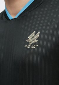 Μαύρο πουκάμισο με λεπτές κάθετες ρίγες, γιακά με μπλε περίγραμμα και ασημένιο λογότυπο στο στήθος που γράφει "Milano Italia Est 2004".