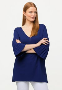 Donkerblauwe top met een V-hals en brede, geplooide mouwen; gemaakt van een geweven stof. Het ontwerp is losjes passend en heuplang.