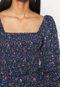 Mustaa kangasta, jossa on kukkakuosi, jossa on sinisiä ja vaaleanpunaisia kukkia. Neliömäinen kaula-aukko, puffihihat ja kerrostettua tekstuuria kauttaaltaan.