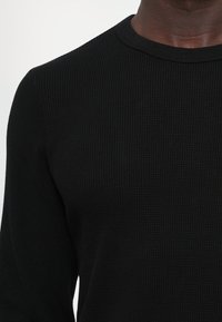 Close-up of a person wearing a black textured long-sleeve top with a round neckline against a plain light background.