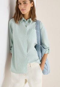 Femme portant une chemise à rayures vert clair à boutons et un pantalon blanc, avec un sac fourre-tout bleu clair sur l'épaule.