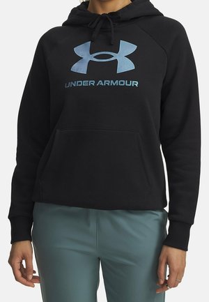 Person trägt einen schwarzen Under Armour Hoodie mit blauem Logo und Text, kombiniert mit blau-grünen Hosen, steht mit entspannten Armen.