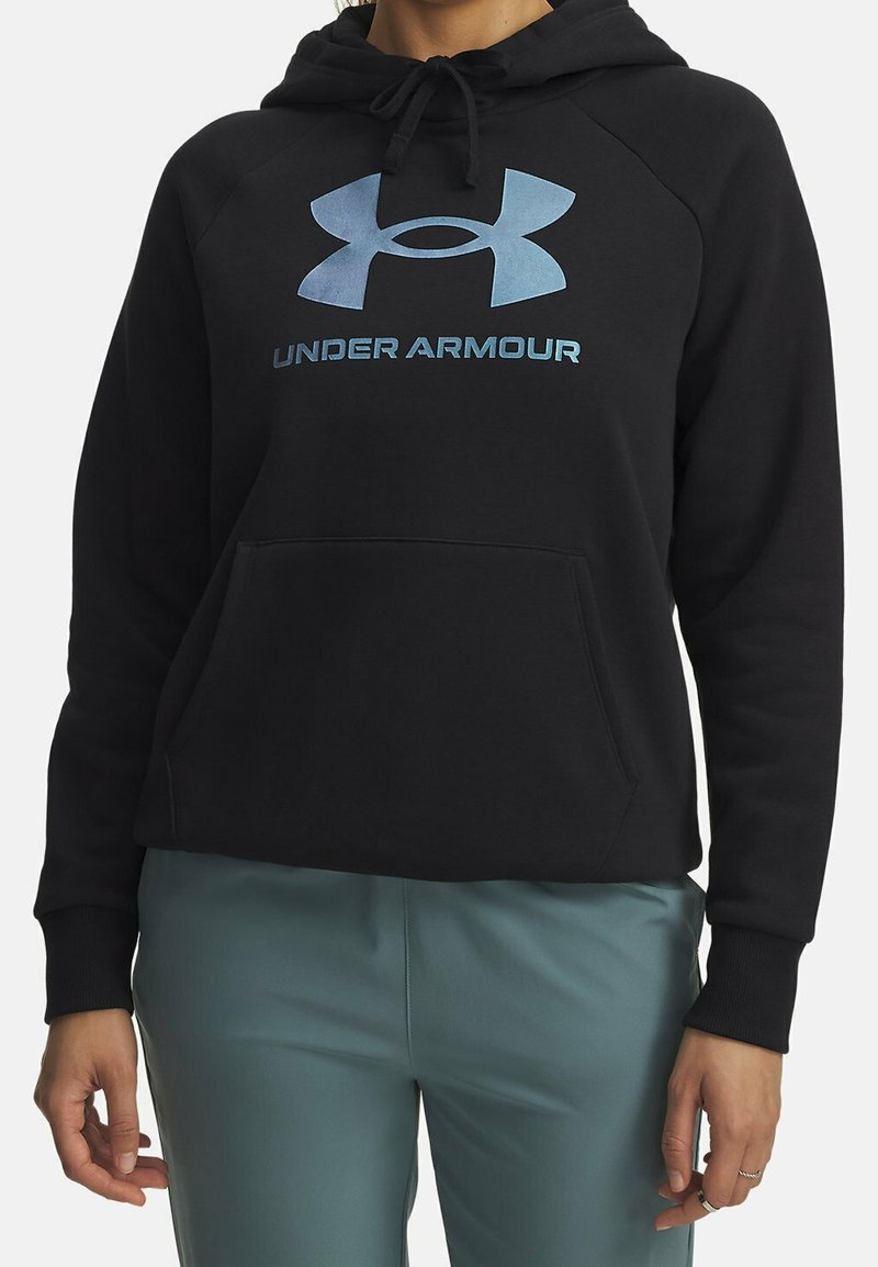 Personne portant un sweat à capuche noir Under Armour avec un logo bleu et du texte, associé à un pantalon bleu-vert, debout les bras détendus.