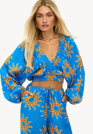 Blauwe cropped top en bijpassende broek met een zonnenpatroon in oranje. Heeft puffed sleeves, een diepe V-hals en een elastische tailleband.