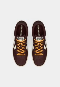Zapatillas Converse All Star en ante burdeos oscuro con detalles en crema, presentan cordones naranjas y una suela de goma texturizada.