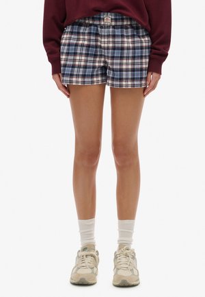 Person iført blå og hvide ternede shorts, hvide sokker, beige sneakers og en langærmet vinrød top, vist fra midt på låret og ned.