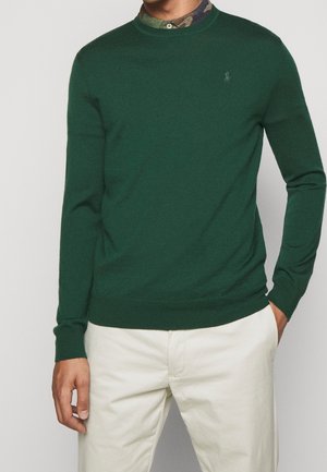 Mann trägt einen dunkelgrünen Pullover über einem Tarnmuster-Hemd mit cremefarbenen Hosen, Hand in der Tasche, steht vor einem schlichten Hintergrund.