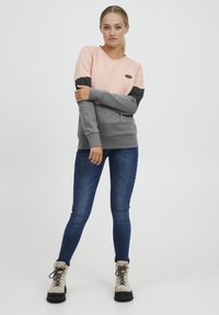 Sweatshirt rose clair et gris foncé avec un col rond, des manches longues et une petite pièce de logo. Associé à un jean skinny bleu et des bottes beiges.