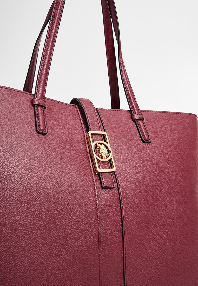 Bolso tote de cuero burdeos con textura suave, que presenta asas alargadas y un cierre de hardware en tono dorado con un emblema decorativo.