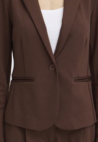 Hnedé priliehavé sako so zapínaním na jeden gombík, dvoma prednými vreckami s rozparkami, nosené cez biely top.