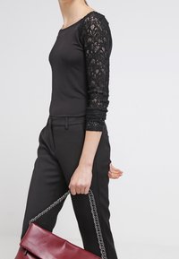 Černý top s krajkovými dlouhými rukávy, zkombinovaný s černými kalhotami a držící burgundskou kabelku s řetízkovým popruhem.