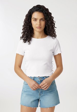 Giovane donna con capelli ricci che indossa una maglietta bianca a maniche corte e pantaloncini di jeans azzurri, in piedi con le mani intrecciate.