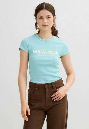 Mujer joven con cabello castaño, vestida con una camiseta azul clara con la inscripción "Nueva York" y pantalones marrones de talle alto, de pie frente a un fondo liso.