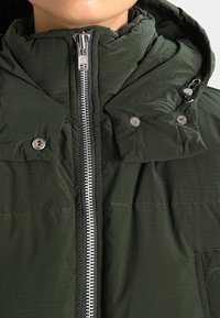 Kurtka puchowa w kolorze oliwkowej zieleni z zamkiem błyskawicznym z przodu, zatrzaskami na kołnierzu oraz teksturowaną, wodoodporną tkaniną. Posiada kaptur.