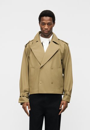 Man draagt een beige cropped trenchcoat met dubbele rij knopen en met knopen gesloten manchetten, gecombineerd met een zwarte broek, staand tegen een effen achtergrond.