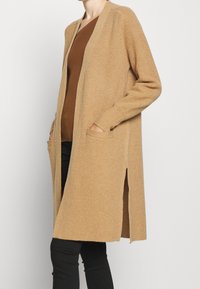 Stickad, lång beige kofta med öppen framsida, ribbad textur, sidoöppningar och två framfickor; matchad med en brun topp och svarta byxor.