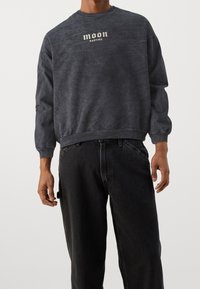 Sudadera de color gris oscuro con textura, que presenta un cuello redondo y puños acanalados, con un logo impreso en blanco que dice "moon KAOTIKO" en el frente.