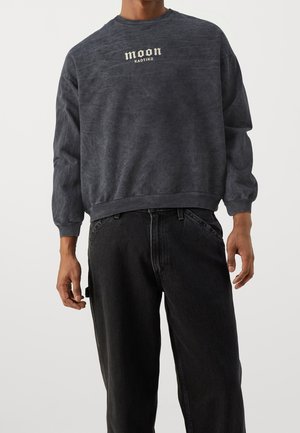 Mörkgrå sweatshirt med struktur, rund halsringning och ribbade ärmar, med en vit tryckt logotyp som säger "moon KAOTIKO" på framsidan.