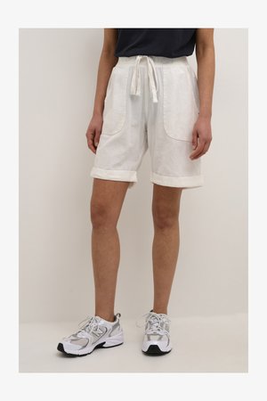 Pantalones cortos de algodón blancos con cintura ajustable, puños enrollados y grandes bolsillos frontales, combinados con zapatillas deportivas grises y blancas.