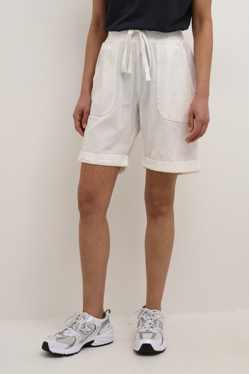 Pantalones cortos de algodón blancos con cintura ajustable, puños enrollados y grandes bolsillos frontales, combinados con zapatillas deportivas grises y blancas.