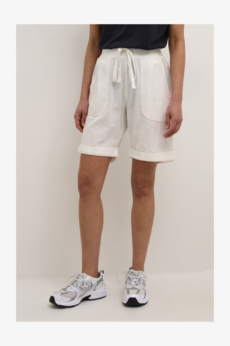 Pantalones cortos de algodón blancos con cintura ajustable, puños enrollados y grandes bolsillos frontales, combinados con zapatillas deportivas grises y blancas.