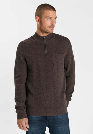 Uomo che indossa un maglione marrone con zip al quarto e tasca sul petto e jeans blu, guarda verso destra su uno sfondo bianco uniforme.