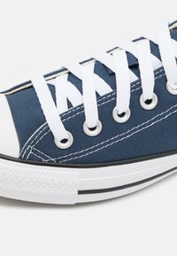 Navy canvas sneaker with white laces, circular eyelets, rubber tip, and black stripe on a white sole. Textured fabric surface detail.