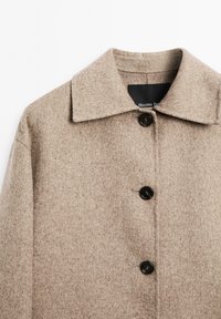 Beige uldfrakke med bred krave og tre sorte knapper foran, der viser den øverste del af brystet og skuldrene.