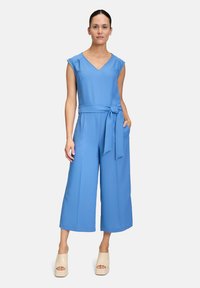 Blauer Jumpsuit mit V-Ausschnitt, kurzen Ärmel, einem gebundenen G�ürtel in der Taille, weiter Beinführung und Taschen. Kombiniert mit beigen Plateausandalen.