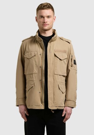 Ung mand iført en beige militærinspireret jakke med flere lommer over en sort skjorte, stående foran en ensfarvet grå baggrund.