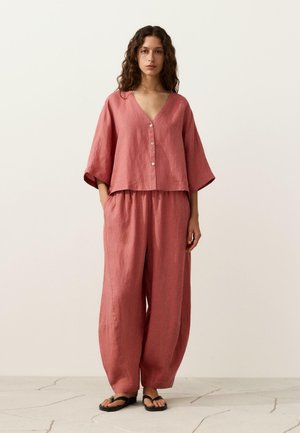 Vrouw met krullend haar die een los roze overhemd met knopen en wijde broek draagt, blootsvoets staand op een lichte tegelvloer.