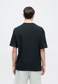 Μαύρο βαμβακερό T-shirt με κοντά μανίκια και στρογγυλή λαιμόκοψη. Η πίσω όψη αποκαλύπτει ένα απλό σχέδιο χωρίς μοτίβα ή γραφικά.