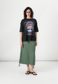 Black graphic t-shirt with multicolored design, paired with a mid-length green skirt and black flip-flops. Features short sleeves and a relaxed fit.
