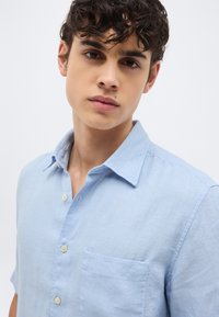 Νεαρός άνδρας με κυματιστά σκούρα μαλλιά, ντυμένος με γαλάζιο πουκάμισο με κουμπιά και τσέπη στο στήθος, κοιτάζει ήρεμα την κάμερα μπροστά σε ένα απλό φόντο.