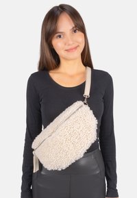 Cremefarbene Crossbody-Tasche aus Kunstfell mit Reißverschluss und flachem Tragegurt. Die strukturierte Oberfläche kontrastiert mit dem glatten Lederimitat-Boden.