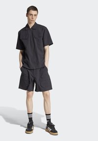 Čierna krátkotrupá zipsová košeľa s dvoma prednými vreckami, spárovaná s cargo šortkami so bočnými vreckami, nosená s čiernymi teniskami a pruhovanými ponožkami.