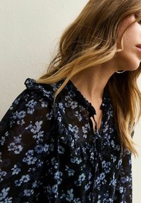 Vrouw met lichtbruin haar, gekleed in een doorschijnende zwarte blouse met blauw bloemmotief en een gerimpelde kraag, tegen een effen achtergrond.