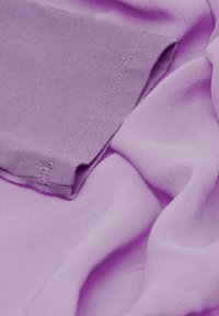 Helllila Stoff mit glatten und strukturierten Lagen, versehen mit sichtbaren Nähdetails an einem gefalteten Abschnitt.