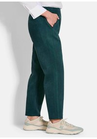 Donkergroene suede broek met zijzakken, taps toelopende pijpen en zichtbare stiksels, gecombineerd met lichtbeige sportschoenen.