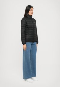 Zwarte pufferjack met hoge kraag, ritssluiting en gewatteerd patroon. Gecombineerd met ruime blauwe jeans en witte sneakers.
