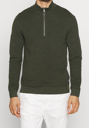 Mężczyzna ubrany w oliwkowozielony prążkowany sweter z krótkim srebrnym zamkiem błyskawicznym i białe spodnie, stojący na jednolitym tle.