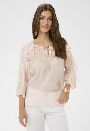 Mladá žena s dlhými hnedými vlasmi, oblečená v svetloružovej blúzke s volánmi a kvetinovým vzorom, biele nohavice, ružové náušnice a zlatý náramok.