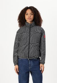Juodai balta zip-up striukė su pakartotiniu teksto raštu, aukštu apykakle ir elastingomis rankovėmis. Derinama su mėlynomis džinsinėmis kelnėmis.